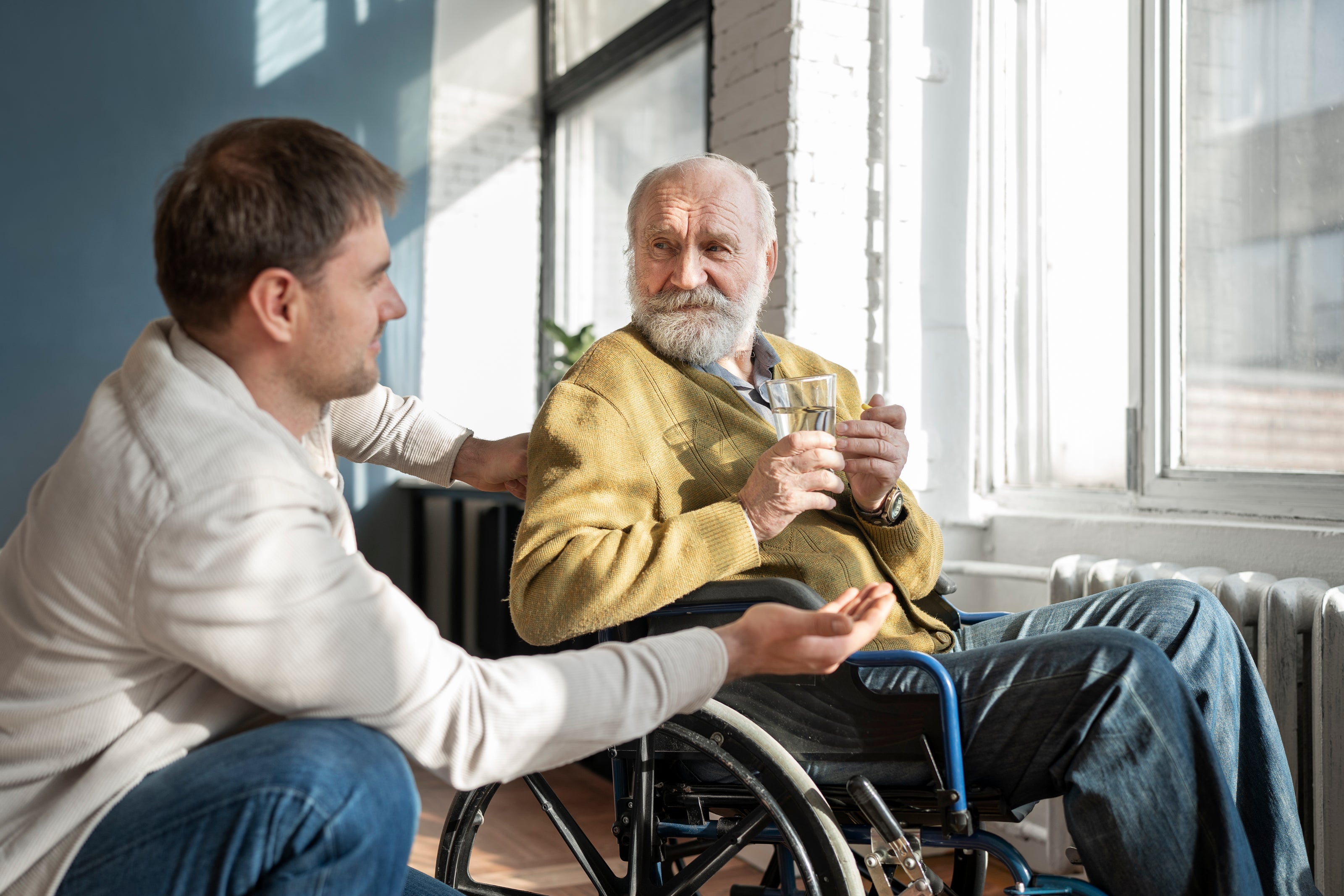 Man in a wheelchair sitting next to an older man with a beard, both by a window.